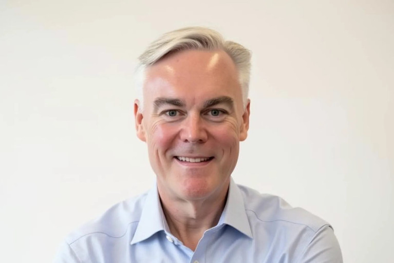Arms crossed and smiling, Tom Christenson stands in front of a white background, wearing a light blue button-down shirt.