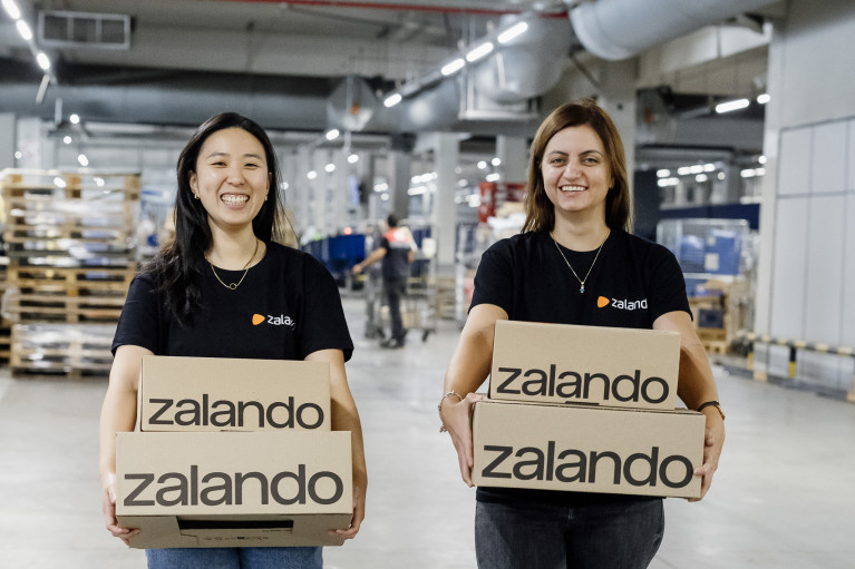 Vivian and Iva, each carrying two Zalando parcels, are walking together in a big warehouse smiling at the camera