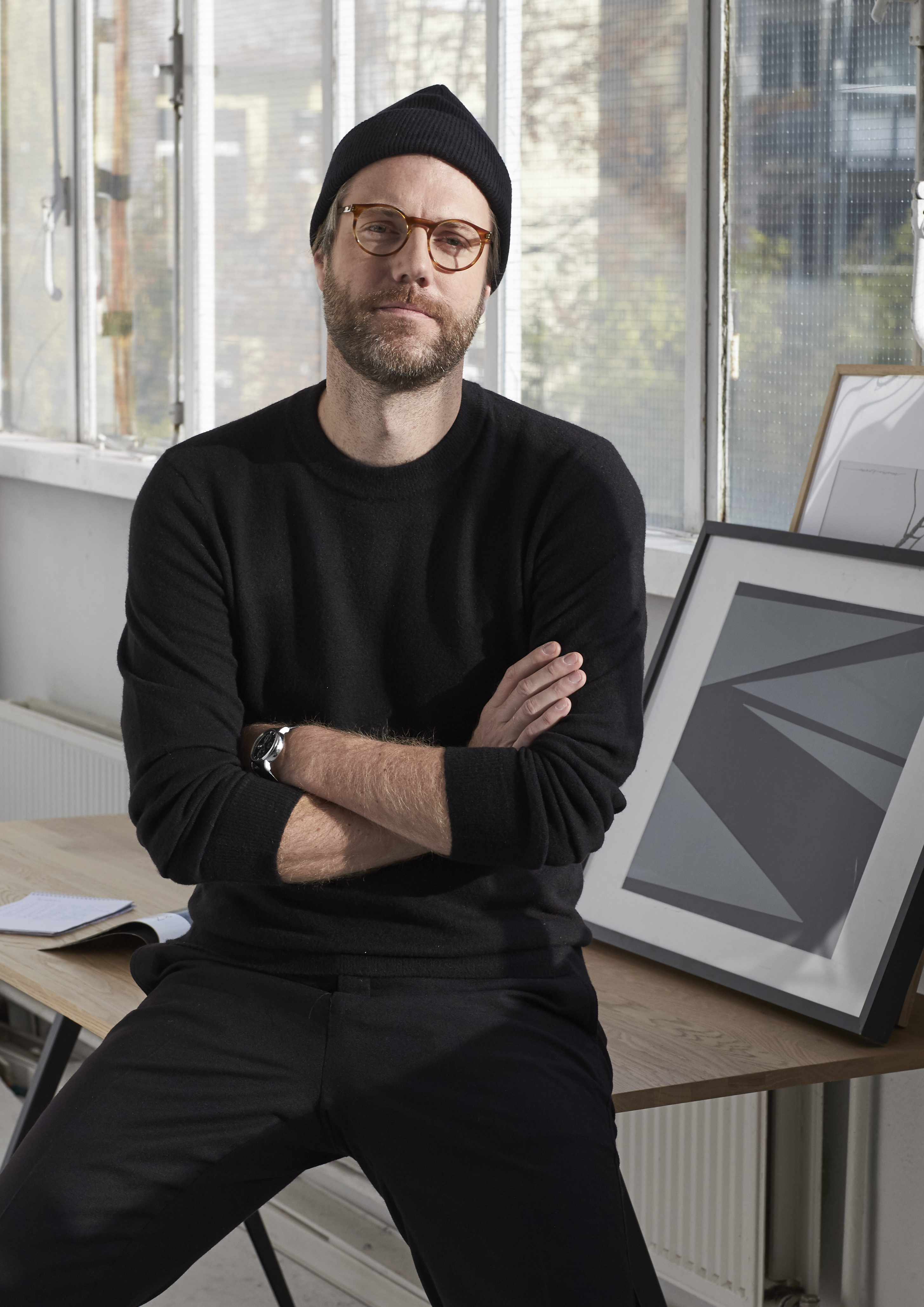 Portrait of Torsten Orendt in glasses and beanie sitting on a desk in front of a big glass window