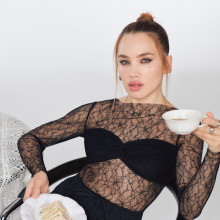 Isamaya Ffrench in a black, partly see-through dress holding a plate with a piece of cake and a white porcelaincup