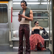 Two people in the underground; a man with headphones is standing up looking at his phone, the woman dressed up in a frilly pink and dark eyeglasses is sitting in the backgroundskirt