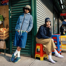 Streetcorner scene next to a greengrocer's with produce displayed outside: a man with sunglasses is relaxedly leaning against the wall, a woman sitting on stacked colourful chairs looking coolly into the camera