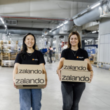 Vivian and Iva, each carrying two Zalando parcels, are walking together in a big warehouse smiling at the camera