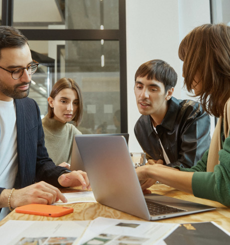 A group of diverse people sitting around a table discussing something on a laptop