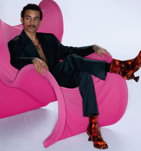 Fashion model sitting on a futuristic pink armchair wearing a black suit and characteristic boots