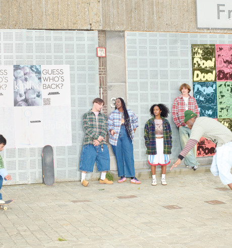 street corner scene: two young men skateboarding while other people stand along a wall watching