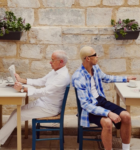 Two men sitting back to back at coffee house tables; the older one reading a newspaper, the younger wearing sunglasses