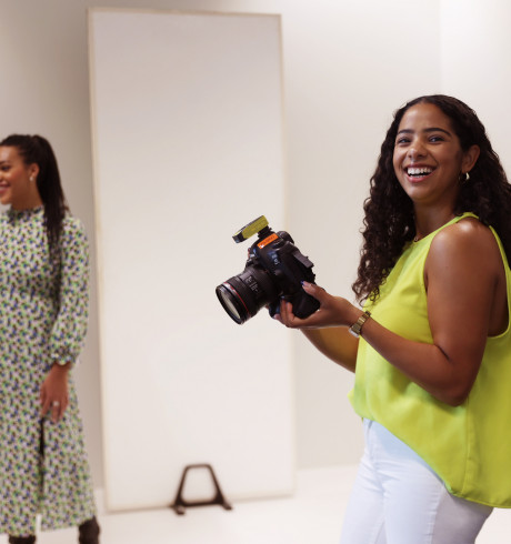 Set of a photoshoot: black female photographer smiling at the camera, black model in the background