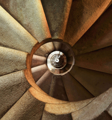 A close-up, downward view of a spiral staircase made of stone. The staircase curves tightly, creating a hypnotic, circular pattern that draws the eye to the center.