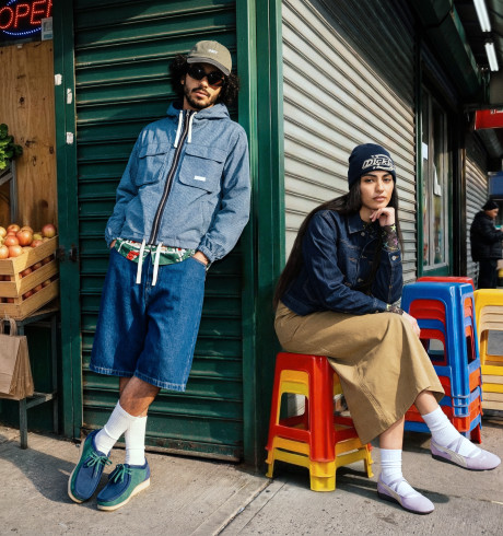Streetcorner scene next to a greengrocer's with produce displayed outside: a man with sunglasses is relaxedly leaning against the wall, a woman sitting on stacked colourful chairs looking coolly into the camera