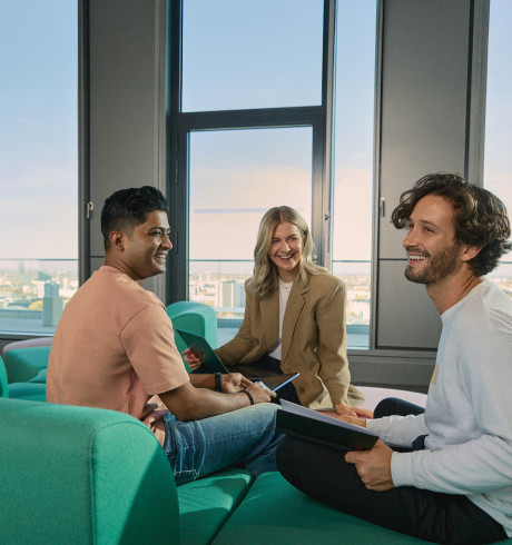 A group of three young professionals sits in a sunlit lounge area, engaged in a cheerful discussion. They are holding tablets and notebooks, suggesting a collaborative meeting. The background features large windows overlooking the city of Berlin, highlighting a modern and open workspace environment.