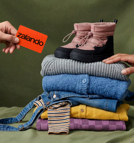 Ein Stapel bunter Kleidung; darauf ein Paar Kinderschuhe; von links kommt eine Hand mit einer Zalando-Karte