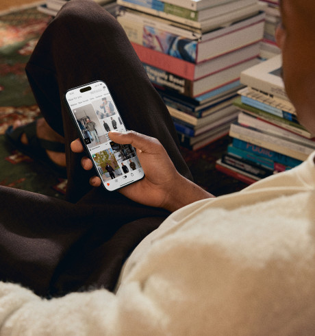 A black man sitting on the floor on an oriental carpet next to piles of books; browsing the Zalando shop on his phone
