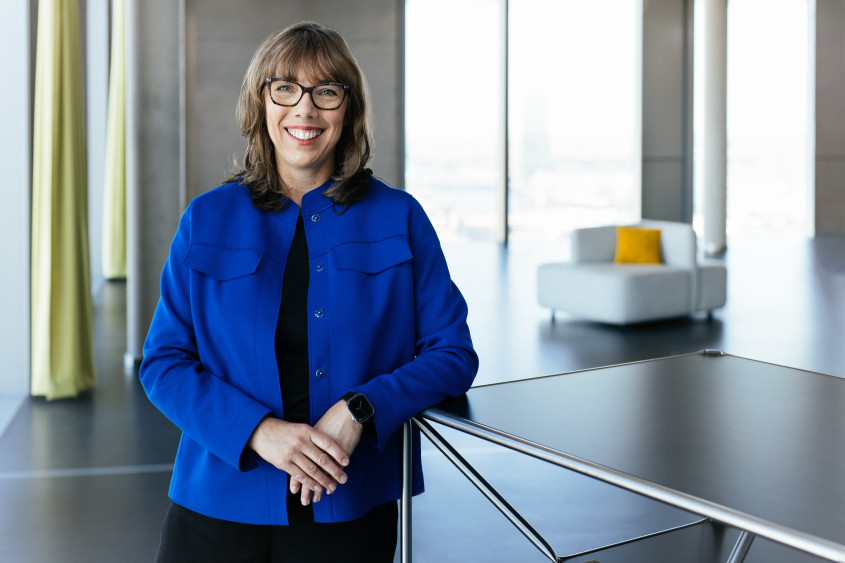 Portrait of Dr. Astrid Arndt: A smiling woman wearing glasses, medium-length brown hair, and a royal blue jacket leans against a metal table in a modern, light-filled office building.