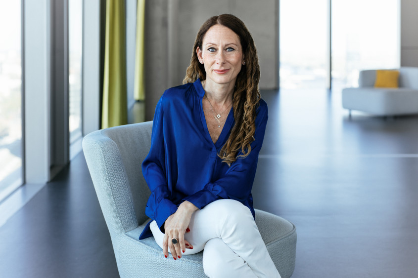 Portrait of Anna Dimitrova: A woman with long, slightly wavy brown hair seated on a light grey armchair. She wears a blue silk blouse, white trousers, and smiles into the camera.