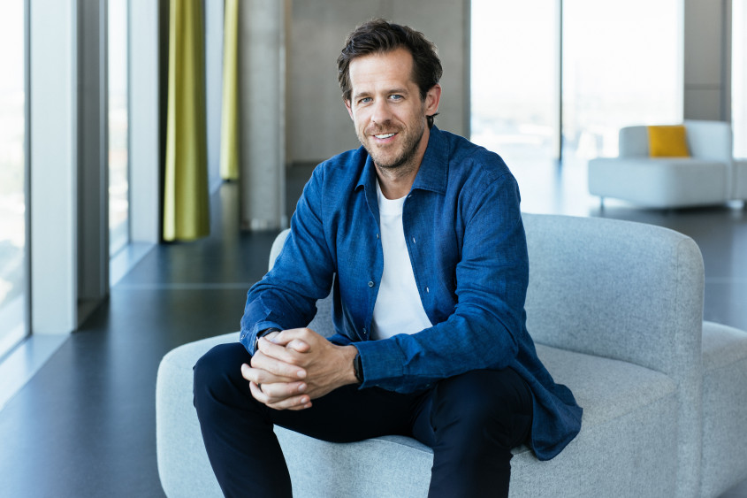 Portrait of Robert Gentz: A smiling man with short brown hair sits on a light grey sofa. He wears a blue shirt over a white T-shirt and dark trousers.