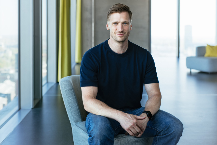 Portrait of David Schneider: A man with short light brown hair sits smiling on a grey armchair. He wears a dark T-shirt, jeans, and looks directly at the camera.