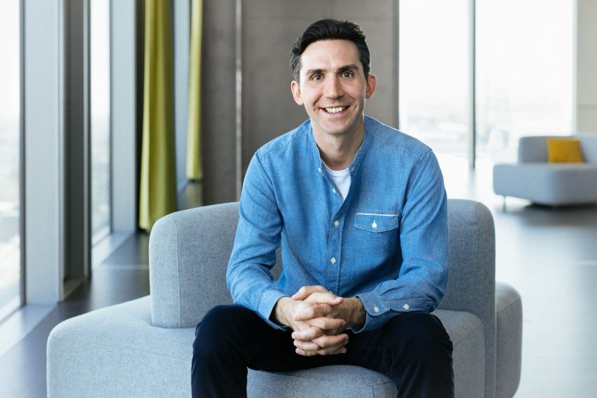Portrait of David Schröder: A smiling man with dark hair sits on a light grey armchair with his hands clasped in his lap. He wears a light blue denim shirt and dark trousers.