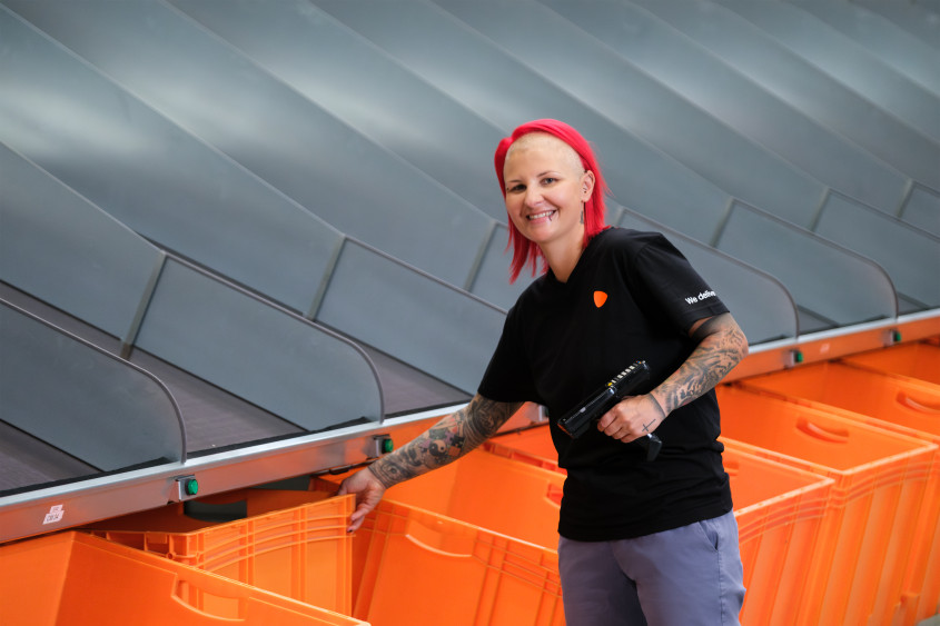 Ein:e Zalando-Mitarbeiter:in mit roten Haaren, Piercings und Tattoos, gekleidet in ein schwarzes Uniformhemd und eine blaue Hose, steht vor mehreren orangefarbenen Plastikboxen.