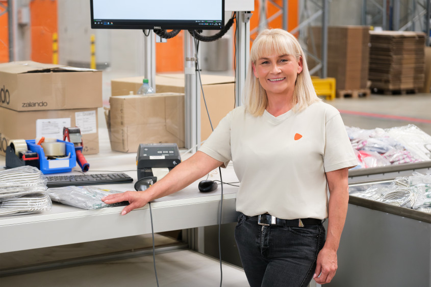 Eine Frau in einem hellen T-Shirt lächelt, während sie an einem ordentlichen Packtisch steht, der mit einem Computermonitor, einem Barcodescanner und Versandetiketten ausgestattet ist.
