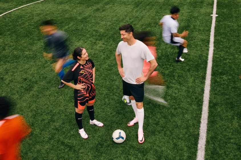 Robert Lewandowski and Alexia Putellas standing on the football field in conversation with a ball between them; around them there are other blurry players in action