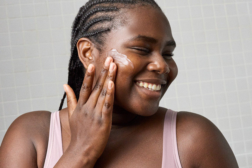 Black woman during beauty routine; wearing only a pink bra, applying product to their face and smiling