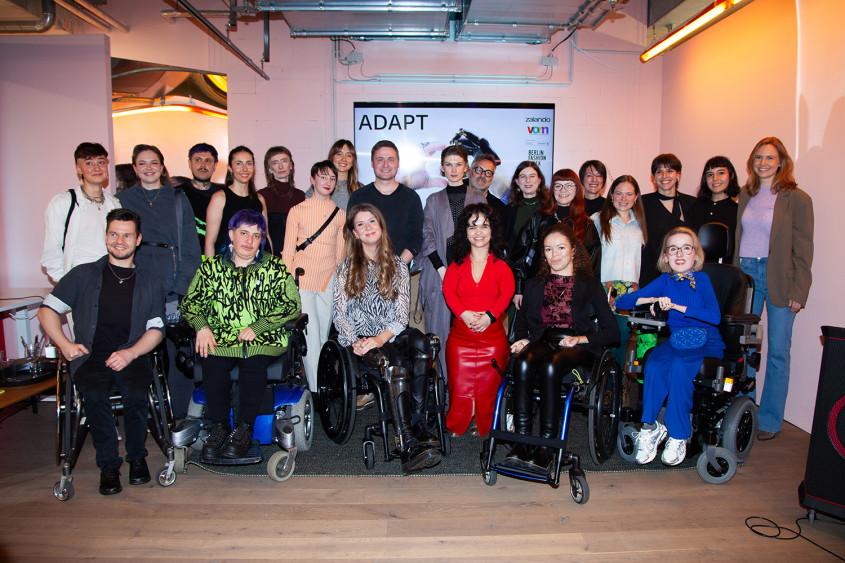 Eine Gruppe von etwa zwölf Personen posiert gemeinsam lächelnd vor einem weißen Banner mit der Aufschrift: vorn x zalando Design academy ADAPT. Die diverse Gruppe umfasst unter anderem Personen im Rollstuhl sowie eine kleinwüchsige Frau in einem auffälligen roten Outfit im vorderen Zentrum.