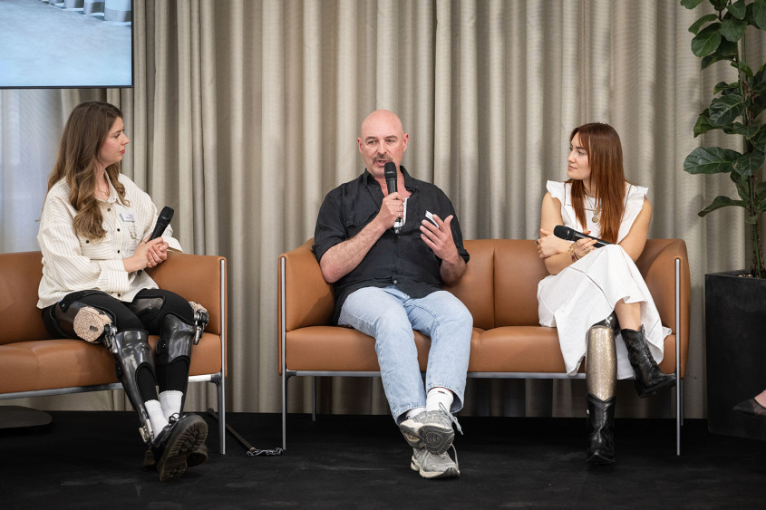 Three panelists sit on brown leather couches on stage holding microphones. The woman on the left wears bilateral leg orthotics, the man in the middle speaks into his mic in a black shirt and light jeans, and the woman on the right sits in a white dress with a visible prosthetic left leg.