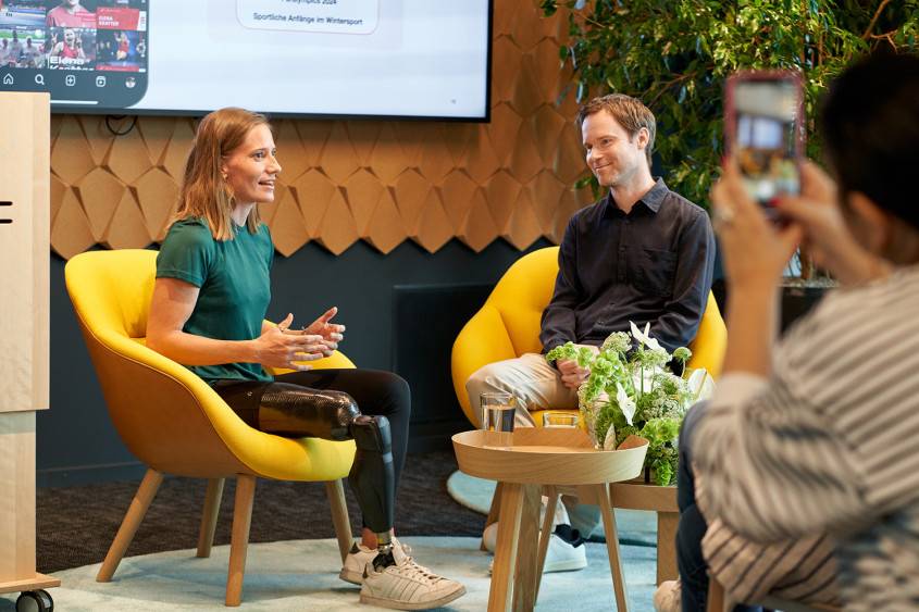 A woman with a prosthetic leg sits in a yellow armchair gesturing while speaking during a presentation. A man listens in a matching yellow chair beside a coffee table with flowers, while an audience member in the foreground captures the moment on a smartphone.