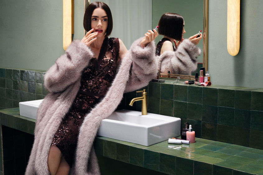 Lily Collins wearing a glamourous sparkly dress and furry coat leaning against a big sink holding her lipstick