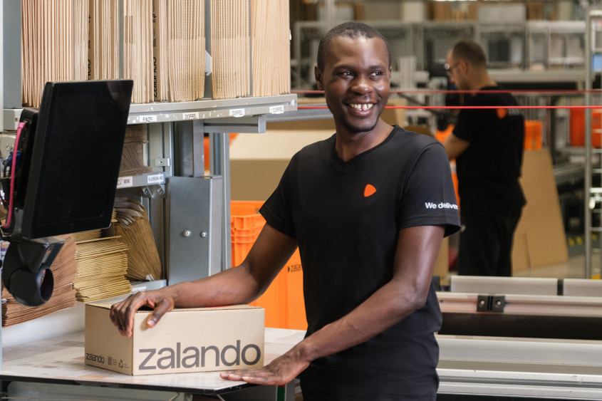 A smiling black man in a black t-shirt and dark cargo pants stands at a warehouse workstation. He is leaning his hands on a cardboard box with the "zalando" logo, surrounded by organized stacks of packaging materials and shipping infrastructure.