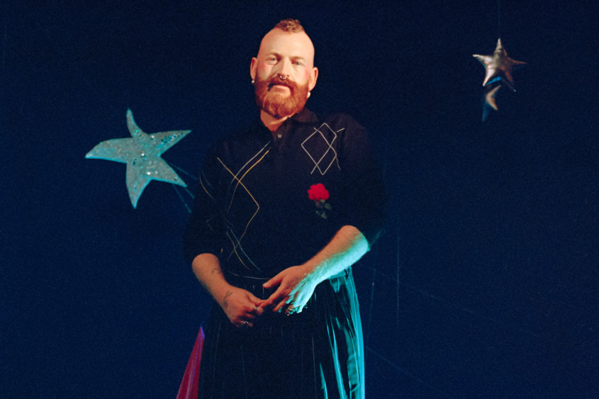 Halbnahaufnahme von Performer Tom Rasmussen mit rötlichem Bart und Irokesenschnitt. Er schaut in die Kamera und trägt ein schwarzes Hemd mit abstraktem Muster und roter Rose sowie einen schwarzen Faltenrock. Seine Hände sind verschränkt. Er steht vor einem dunkelblauen Hintergrund mit hängenden, silbernen Dekosternen.
