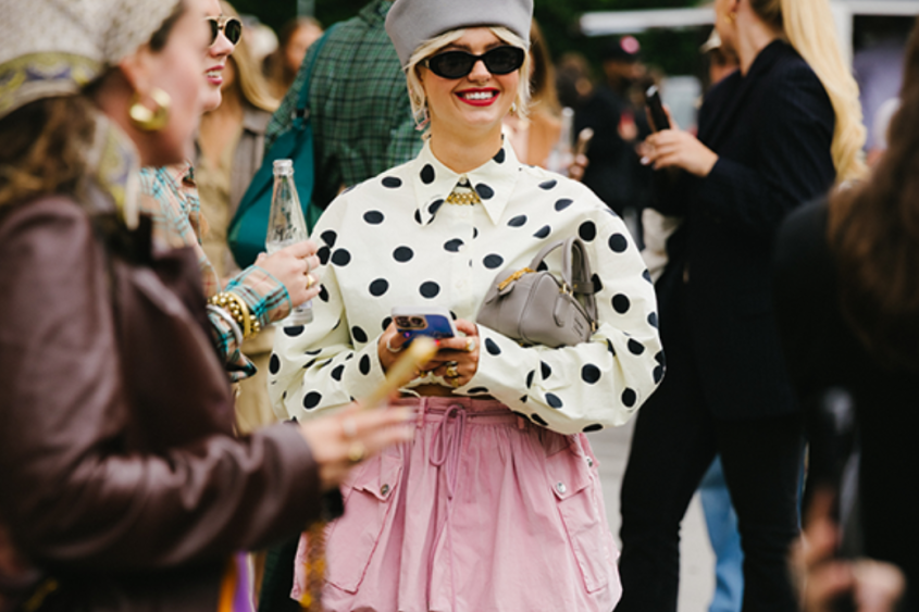 a group of people outdoors, most prominently one young woman in sunglasses a black-and-white polkadot shirt and pink miniskirt