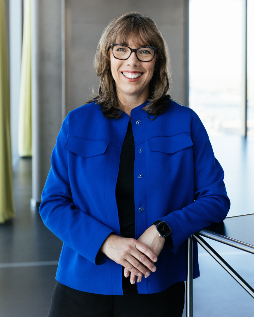 Porträt von Dr. Astrid Arndt: Eine lächelnde Frau mit Brille, mittellangen braunen Haaren und königsblauer Jacke stützt sich in einem modernen, lichtdurchfluteten Bürogebäude an einem Metalltisch ab.