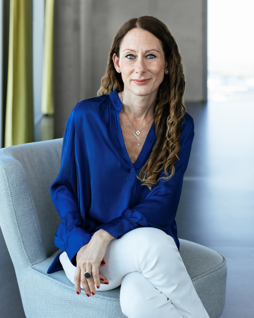 Porträt von Anna Dimitrova: Eine Frau mit langen, leicht gewellten braunen Haaren sitzend auf einem hellgrauen Sessel. Sie trägt eine blau-seidene Bluse, weiße Hosen und lächelt in die Kamera.