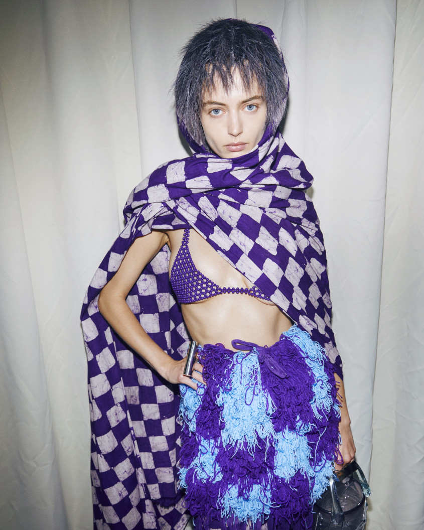 Fashion model backstage wearing a purple bikini top and woolly blue and purple chequered skirt; an oversized white and purple chequered scarf is thrown around their shoulders