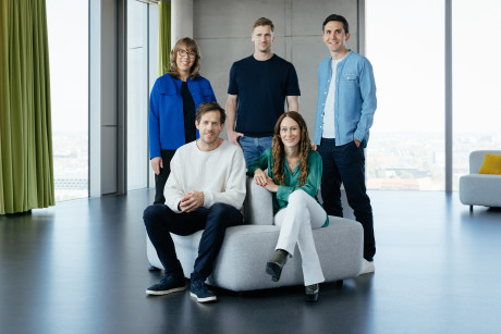Group photo of the Zalando SE Management Board: Five individuals pose together in a modern, bright office space with large windows. Two people are seated in the foreground on grey upholstered furniture, while three others stand behind them.