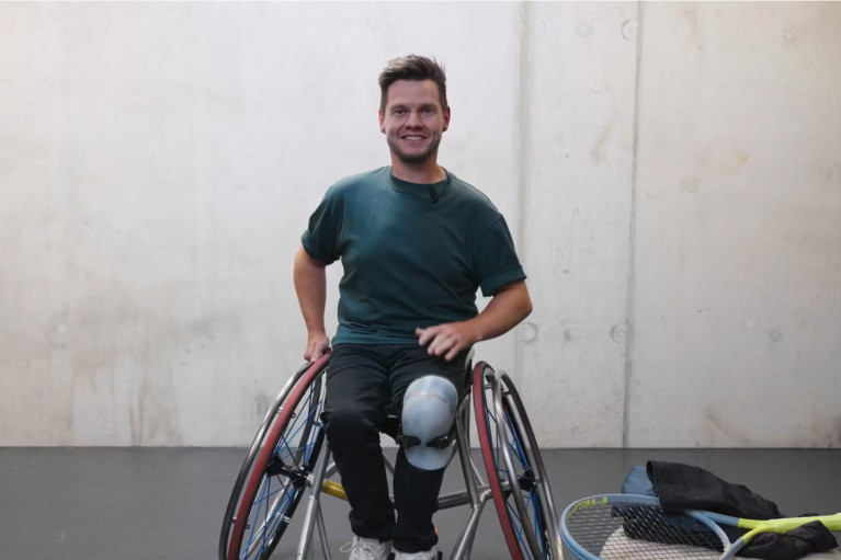 Elite tennis player Marcus Laudan posing for the camera in his wheelchair, on the ground next to him there is a tennis racket and other sports gear
