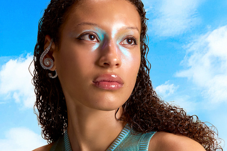 A woman's head and shoulders with shiny makeup and a blue sky and white clouds as background. 
