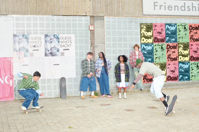 street corner scene: two young men skateboarding while other people stand along a wall watching
