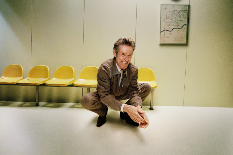 Willem Dafoe dressed in a brown suit crouching in front of a row of yellow chairs