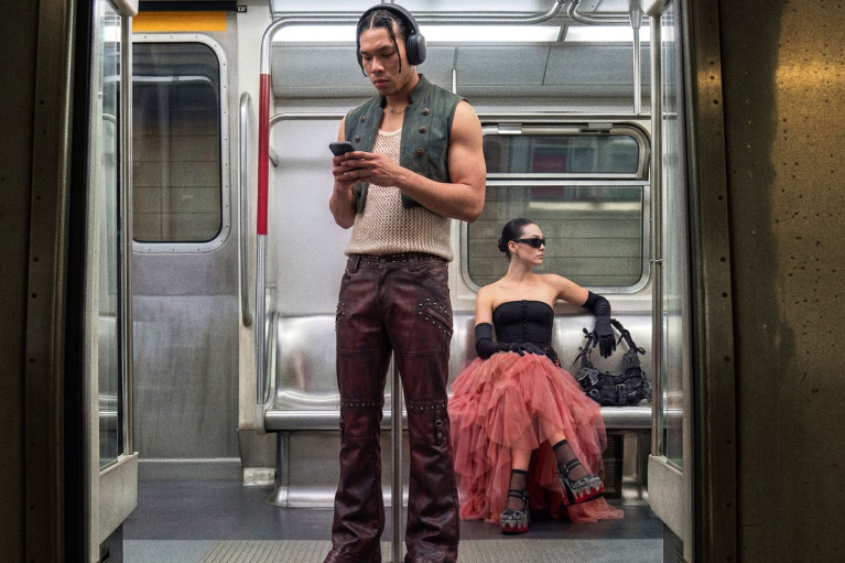 Two people in the underground; a man with headphones is standing up looking at his phone, the woman dressed up in a frilly pink and dark eyeglasses is sitting in the backgroundskirt