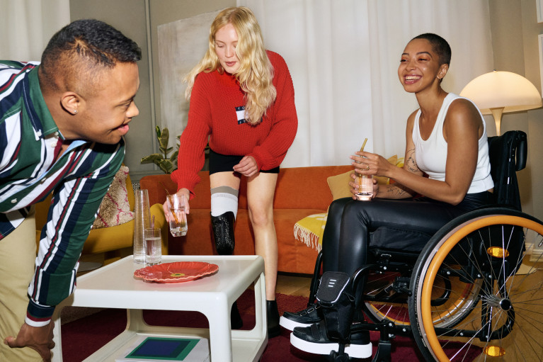 Three interacting young people in a living room scenario; one with a leg brace, one in a wheelchair