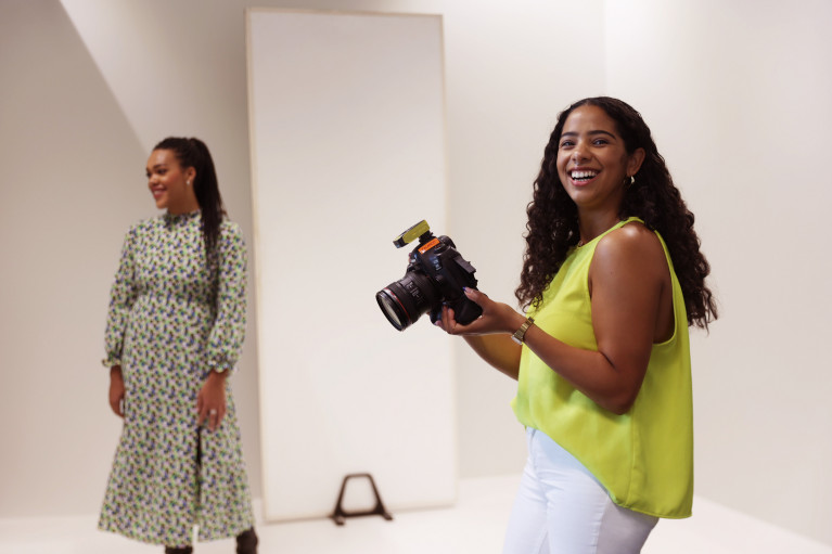 Set of a photoshoot: black female photographer smiling at the camera, black model in the background