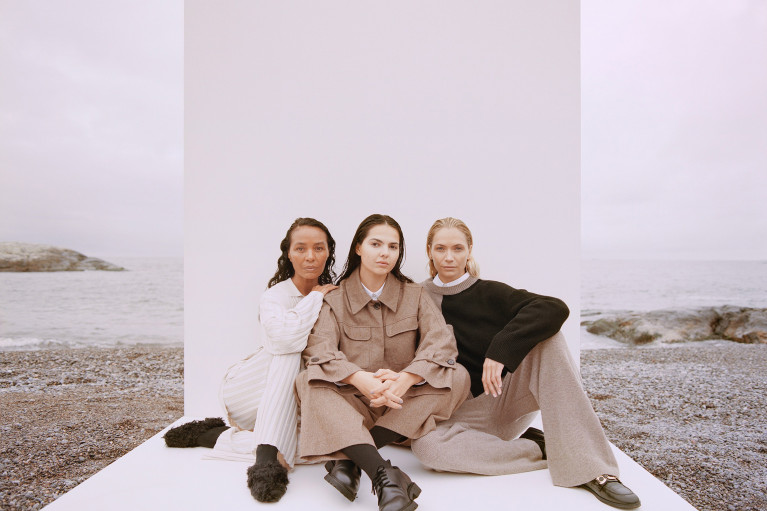 Three fashion models dressed in natural colours sitting in front of a white backdrop surrounded by a foggy beach scenery
