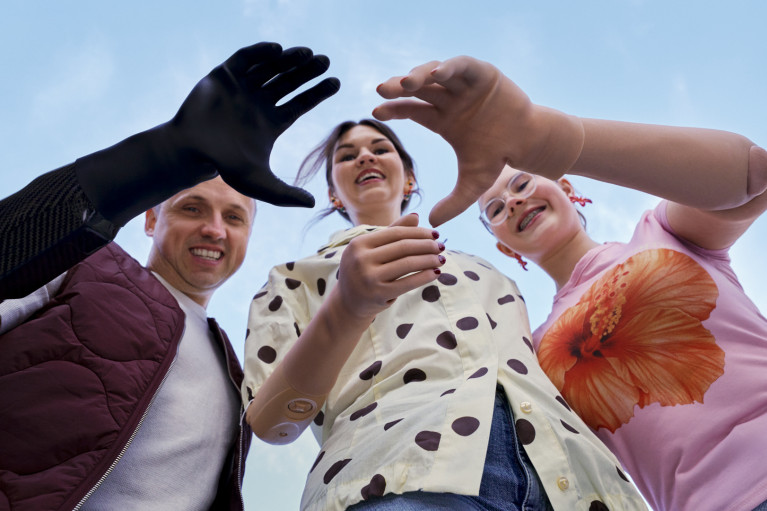 A low-angle shot looking up at three smiling people against a blue sky as they extend their hands and prosthetic arms toward the frame. On the left, a man wears a dark vest and black prosthetic arm; on the right, a girl in glasses and a pink floral t-shirt reaches out with a beige prosthetic arm.