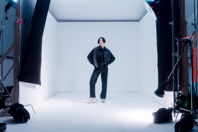 Female fashion model on set; white background, camera equipment in the foreground framing her
