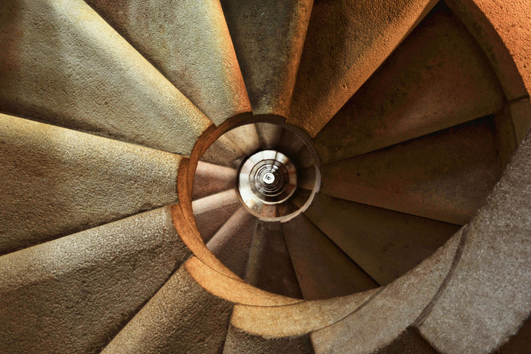 A close-up, downward view of a spiral staircase made of stone. The staircase curves tightly, creating a hypnotic, circular pattern that draws the eye to the center. 