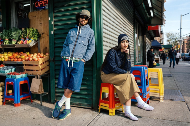 Streetcorner scene next to a greengrocer's with produce displayed outside: a man with sunglasses is relaxedly leaning against the wall, a woman sitting on stacked colourful chairs looking coolly into the camera