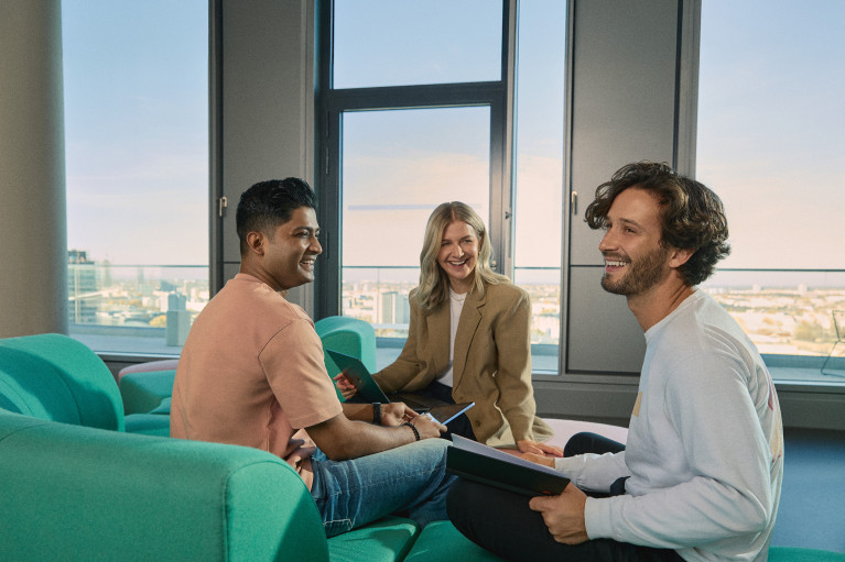 A group of three young professionals sits in a sunlit lounge area, engaged in a cheerful discussion. They are holding tablets and notebooks, suggesting a collaborative meeting. The background features large windows overlooking the city of Berlin, highlighting a modern and open workspace environment.